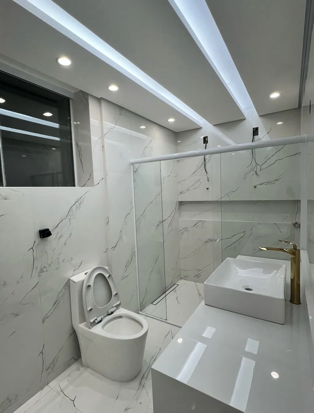 Banheiro de apartamento reformado com revestimento cerâmico marmorizado em tons claros, teto de gesso com sancas iluminadas por LED, bancada em quartzo branco com cuba de sobrepor e torneira monocomando dourada em destaque.
