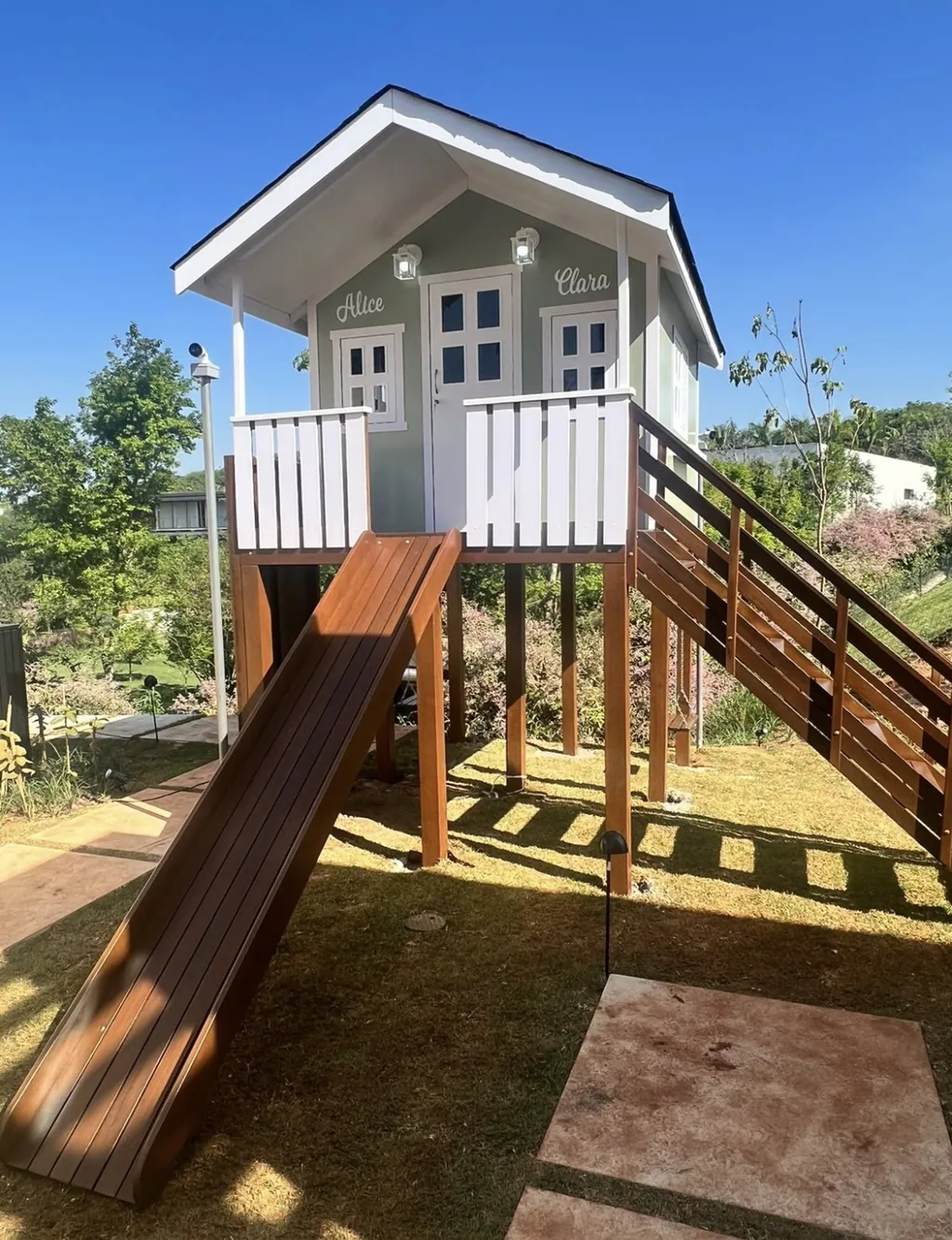 Casinha de brinquedo infantil em madeira sobre pilotis no jardim, pintada em tom verde-oliva claro com detalhes brancos e os nomes Alice e Clara na fachada. Possui escorregador de madeira e escada de acesso.