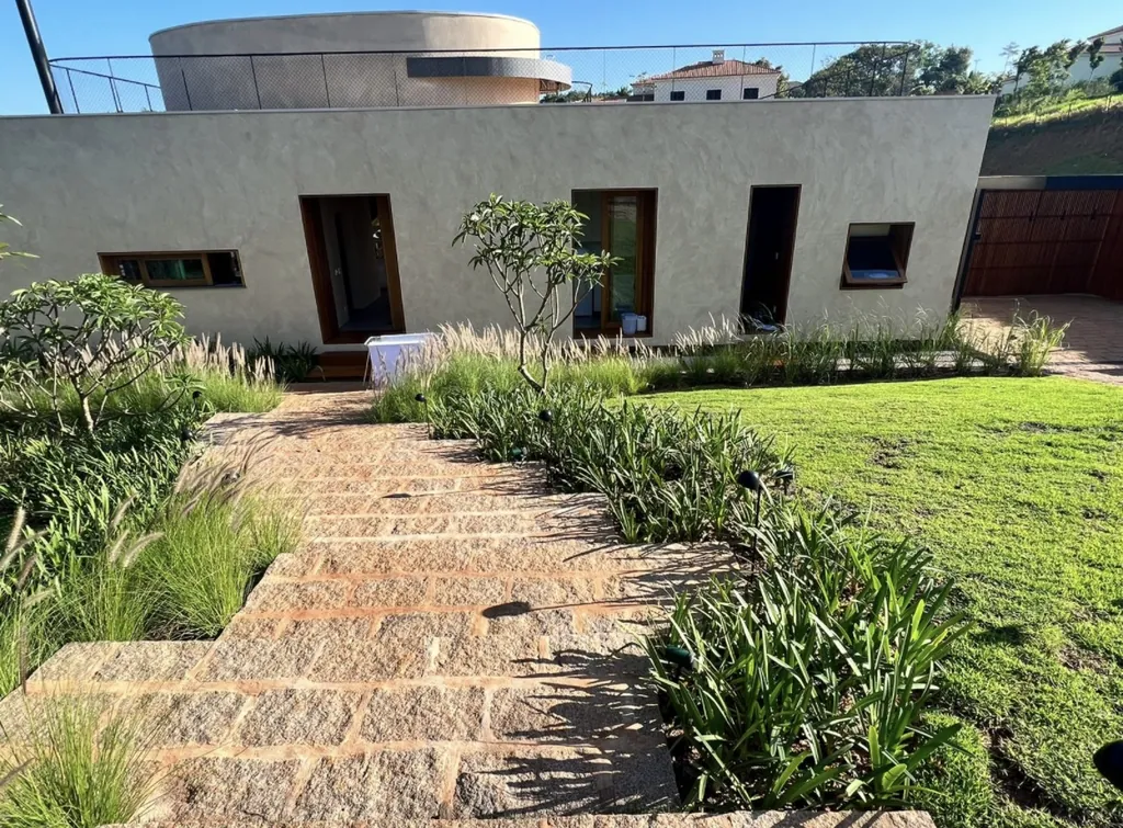 Fachada de casa térrea contemporânea em textura mineral bege com aberturas em caixilhos de madeira. No primeiro plano, escada externa executada em blocos de pedra natural cercada por gramado e capim-dos-pampas.