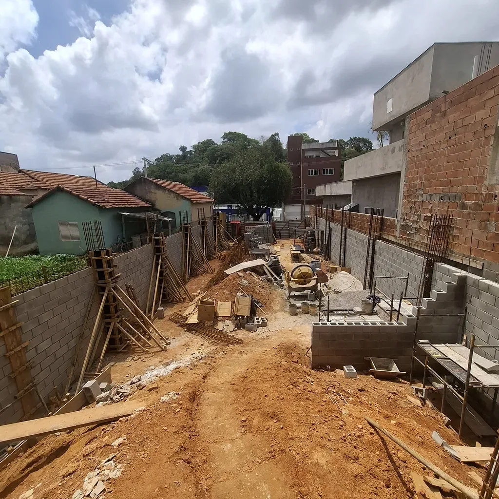 Canteiro de obras externo de galpão comercial em terreno declive com terra vermelha, escoramento de madeira para pilares, paredes de arrimo em blocos de concreto e betoneira ao centro.