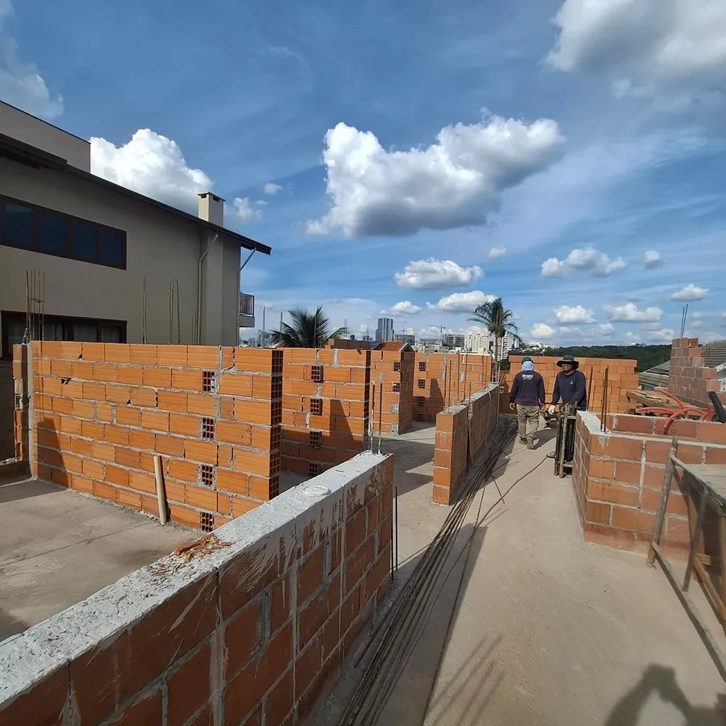Corredor entre paredes de tijolos em execução no pavimento superior, com trabalhadores ao fundo.